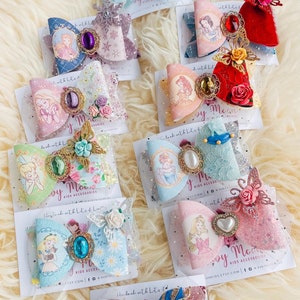 May include: A collection of handmade hair bows featuring various Disney princesses. Each bow is decorated with glitter, lace, and embellishments. The bows are displayed on white cards with the text "Handmade with lots of love" and "By Mommy".