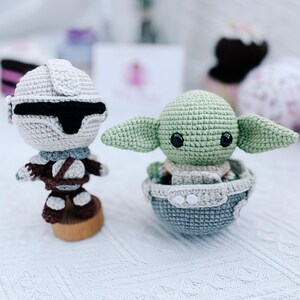 May include: Two crocheted figures: a Stormtrooper and a Grogu (Baby Yoda). The Stormtrooper is in a white helmet and armor, with a black visor. Grogu is green, with large ears, sitting in a gray and blue crocheted pod. Both figures are on a white surface.
