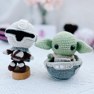 May include: Two crocheted figures: a Stormtrooper and a Grogu (Baby Yoda). The Stormtrooper is in white, gray, and brown, mounted on a wooden base. Grogu is green with large ears, seated in a gray and beige crocheted pod.