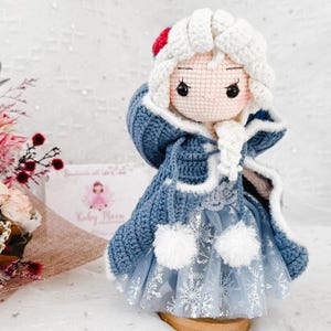 May include: A handmade crochet doll dressed in a blue coat with white trim and a light blue dress with snowflake accents. The doll has white hair and a red accent on the hat. The doll is displayed on a wooden stand.