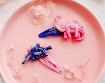 May include: Two dinosaur hair clips on a pink plate. The dinosaur on the left is blue and purple with pink accents. The dinosaur on the right is pink with blue and white accents. Both clips have a blue and pink bar.
