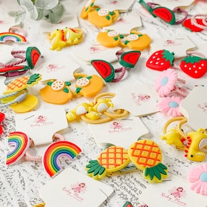 K&ouml;nnte beinhalten: Sortiment an bunten Haargummis mit verschiedenen Obst- und Tiermotiven. Die Haargummis sind auf wei&szlig;en Karten verpackt, auf denen der Text "Ruby Moon" gedruckt ist.