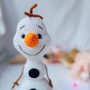 May include: Crocheted snowman toy with a white body, black button accents, and an orange carrot nose. The snowman has a smiling face with black eyes and brown twig arms and hair. A handmade, whimsical character.