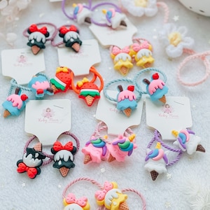 Peut inclure: Assortiment de chouchous en forme de cornet de glace de différentes couleurs et motifs, dont une licorne, une fraise et un Mickey Mouse. Les chouchous sont emballés sur des cartes blanches avec le texte "Ruby Moon" imprimé dessus.