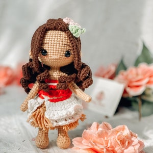 May include: A crocheted doll with brown hair, wearing a white and tan skirt, and a red top. The doll has a flower hair accessory and is posed in front of a soft, blurred background with pink flowers. The doll is handmade.