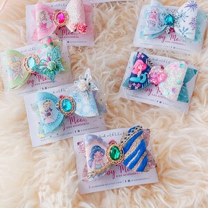 May include: A set of six colorful hair bows featuring various Disney princesses. Each bow is decorated with glitter, sequins, and a jeweled center. The bows are displayed on white cards with the text "Ruby Moon Kids Accessories" and their website address.
