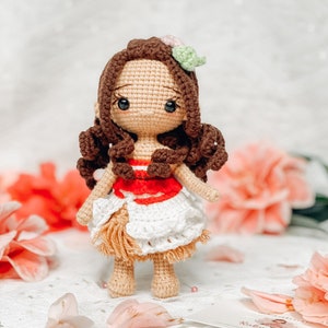 May include: A handmade crochet doll with brown hair, dark eyes, and a floral hair accessory. The doll wears a red and white top and a white skirt with fringe. The doll is standing on a white surface with pink flowers in the background.