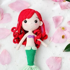 May include: A crocheted mermaid doll with red hair, a green tail, and a purple seashell top. The doll is sitting on a white background with pink flower petals.