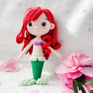 May include: A crocheted mermaid doll with red hair, a purple seashell bra, and a green tail. The doll is sitting on a white surface with pink flower petals.