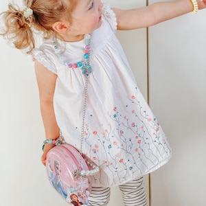 Puede incluir: Una ni&ntilde;a peque&ntilde;a con un vestido blanco con bordado floral, un collar de cuentas y un bolso rosa con un dise&ntilde;o de personaje de Frozen. Tambi&eacute;n lleva una pulsera amarilla y leggings a rayas.