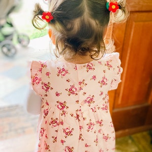 K&ouml;nnte beinhalten: Ein junges M&auml;dchen mit braunen Haaren tr&auml;gt ein rosa Blumenkleid mit einem Knopf im R&uuml;cken. Ihre Haare sind mit roten Blumenhaarklammern nach hinten gezogen.