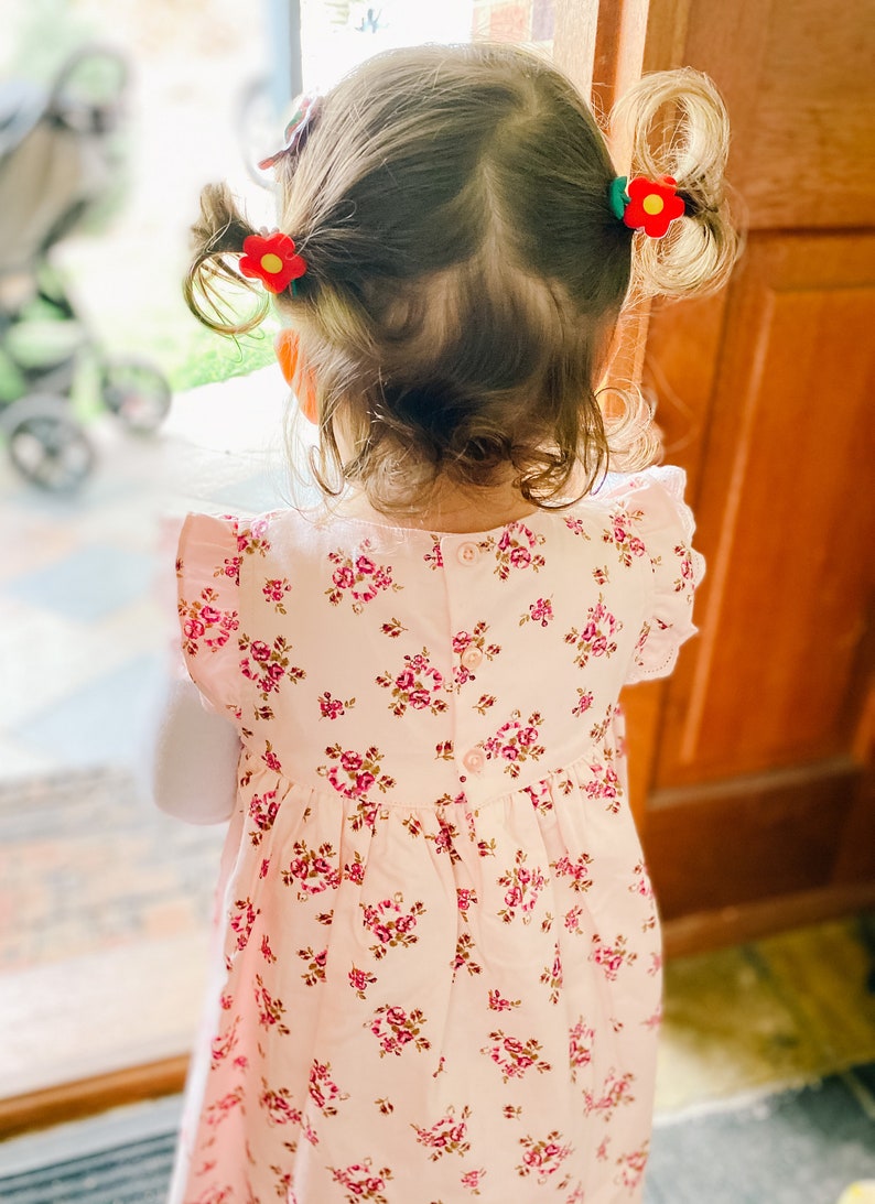 K&ouml;nnte beinhalten: Ein junges M&auml;dchen mit braunen Haaren tr&auml;gt ein rosa Blumenkleid mit einem Knopf im R&uuml;cken. Ihre Haare sind mit roten Blumenhaarklammern nach hinten gezogen.