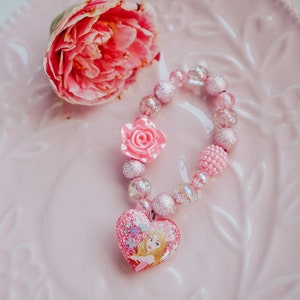 May include: A pink and clear beaded bracelet with a pink flower charm and a heart-shaped charm featuring a princess illustration.