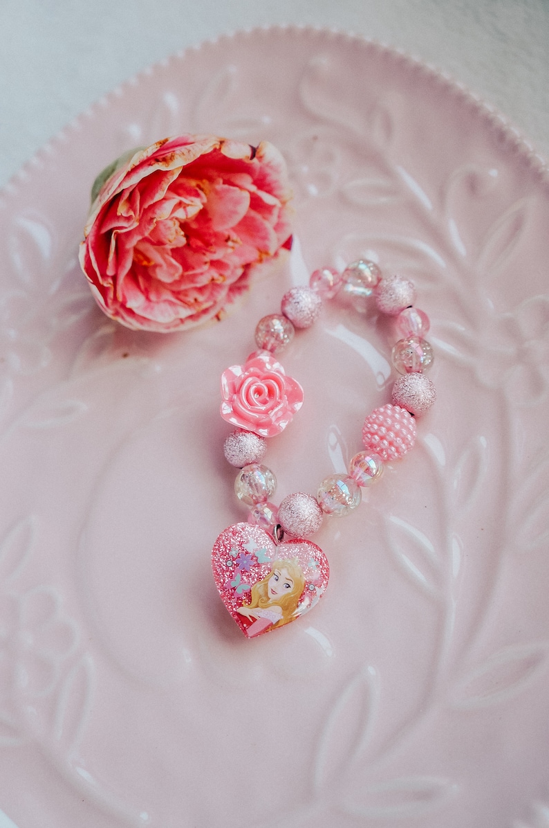 May include: A pink and clear beaded bracelet with a pink flower charm and a heart-shaped charm featuring a princess illustration.