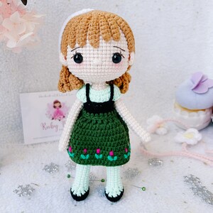 Puede incluir: Una mu&ntilde;eca de crochet con cabello casta&ntilde;o, una diadema blanca, un vestido verde con flores rosas y medias blancas. La mu&ntilde;eca est&aacute; de pie sobre una superficie blanca con copos de nieve plateados.