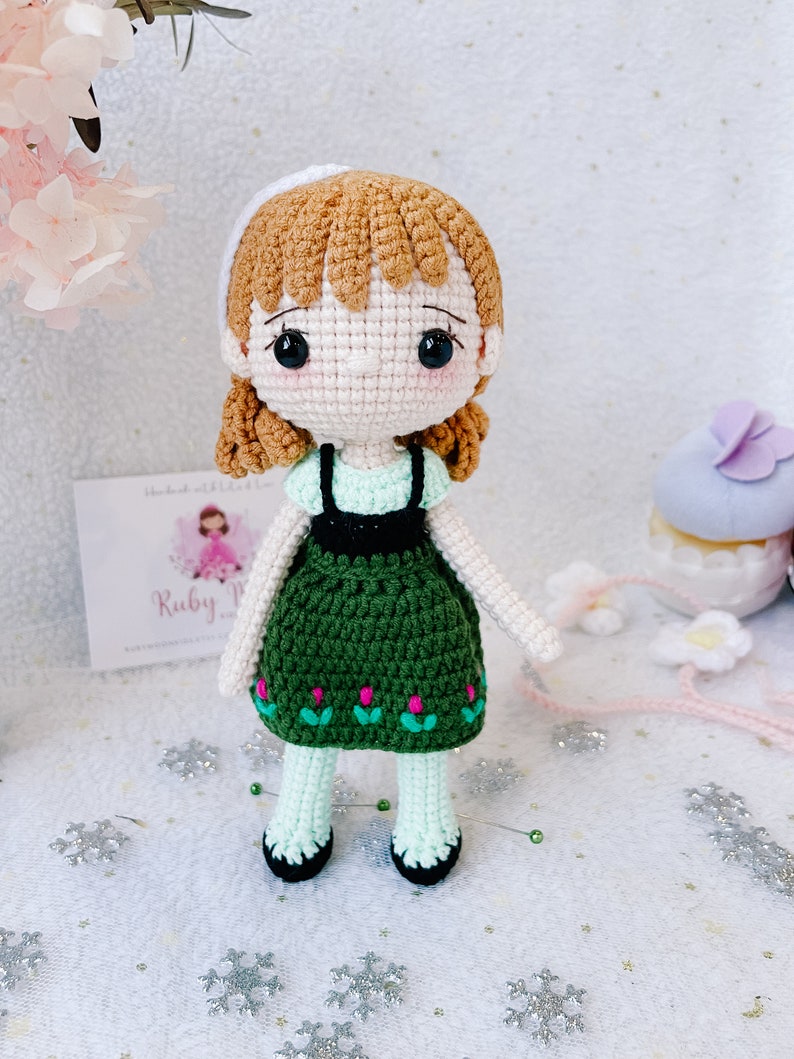 Puede incluir: Una mu&ntilde;eca de crochet con cabello casta&ntilde;o, una diadema blanca, un vestido verde con flores rosas y medias blancas. La mu&ntilde;eca est&aacute; de pie sobre una superficie blanca con copos de nieve plateados.