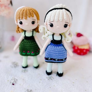 Puede incluir: Dos mu&ntilde;ecas de crochet con el pelo largo, una con el pelo marr&oacute;n y un vestido verde, la otra con el pelo blanco y un vestido azul. Ambas mu&ntilde;ecas tienen ojos negros y llevan zapatos negros.