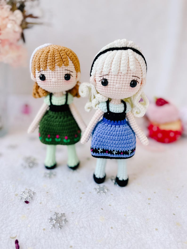 Puede incluir: Dos mu&ntilde;ecas de crochet con el pelo largo, una con el pelo marr&oacute;n y un vestido verde, la otra con el pelo blanco y un vestido azul. Ambas mu&ntilde;ecas tienen ojos negros y llevan zapatos negros.