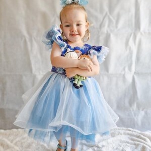 Puede incluir: Una ni&ntilde;a peque&ntilde;a con un vestido de princesa azul y blanco con falda de tul y un lazo azul en el pelo. Est&aacute; sosteniendo dos mu&ntilde;ecas peque&ntilde;as en sus brazos. La ni&ntilde;a est&aacute; de pie sobre una superficie blanca y esponjosa.