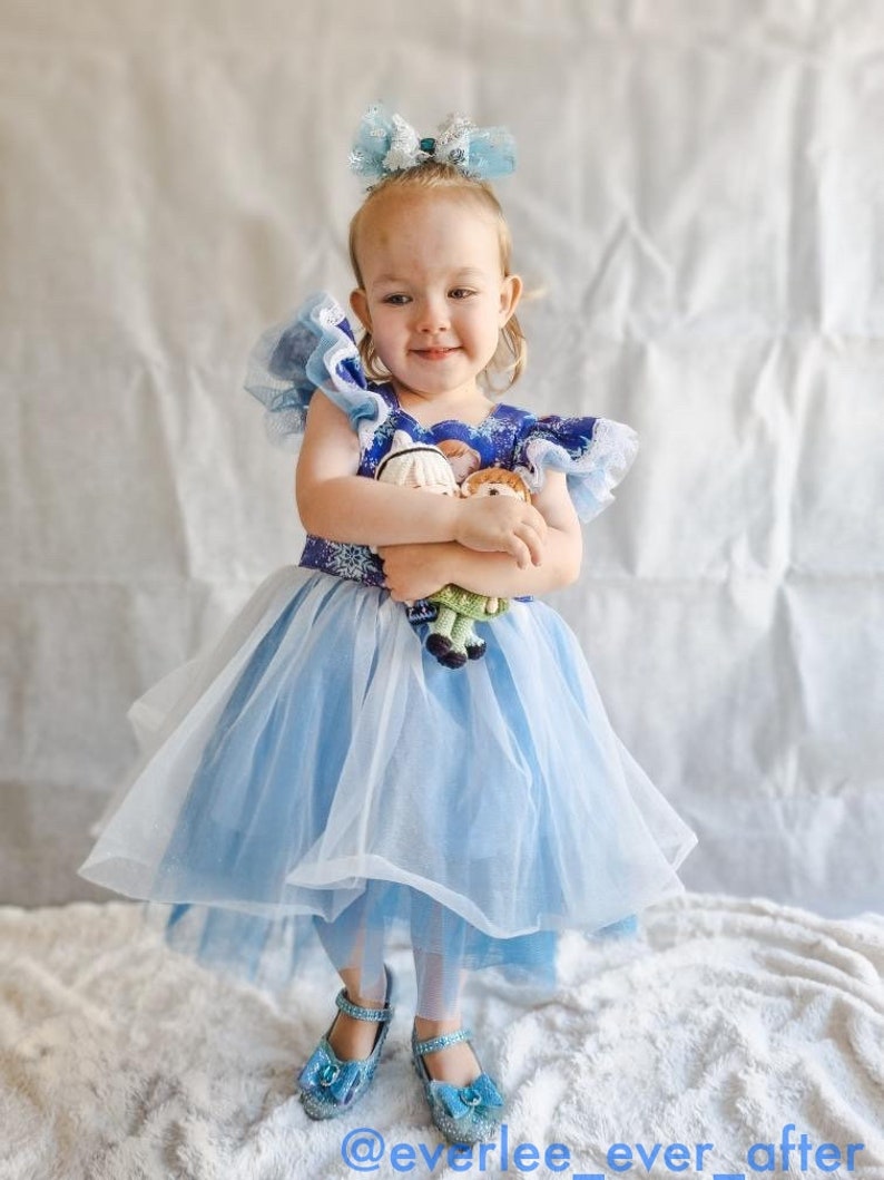 Puede incluir: Una ni&ntilde;a peque&ntilde;a con un vestido de princesa azul y blanco con falda de tul y un lazo azul en el pelo. Est&aacute; sosteniendo dos mu&ntilde;ecas peque&ntilde;as en sus brazos. La ni&ntilde;a est&aacute; de pie sobre una superficie blanca y esponjosa.
