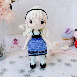 Puede incluir: Una mu&ntilde;eca de crochet con cabello blanco, un vestido azul y zapatos negros. La mu&ntilde;eca est&aacute; de pie sobre un fondo blanco con copos de nieve.