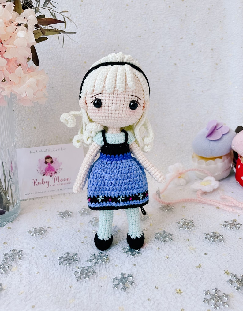 Puede incluir: Una mu&ntilde;eca de crochet con cabello blanco, un vestido azul y zapatos negros. La mu&ntilde;eca est&aacute; de pie sobre un fondo blanco con copos de nieve.
