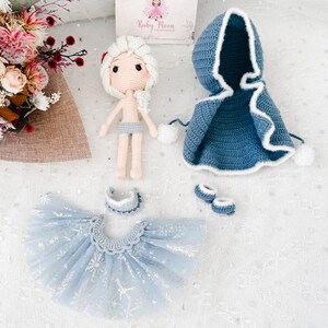 May include: A handmade crochet doll with a white hair and a blue outfit, including a hooded cape, tutu skirt, and shoes. The doll has a light skin tone and is wearing a light blue shorts. The outfit is decorated with white accents and snowflake patterns.