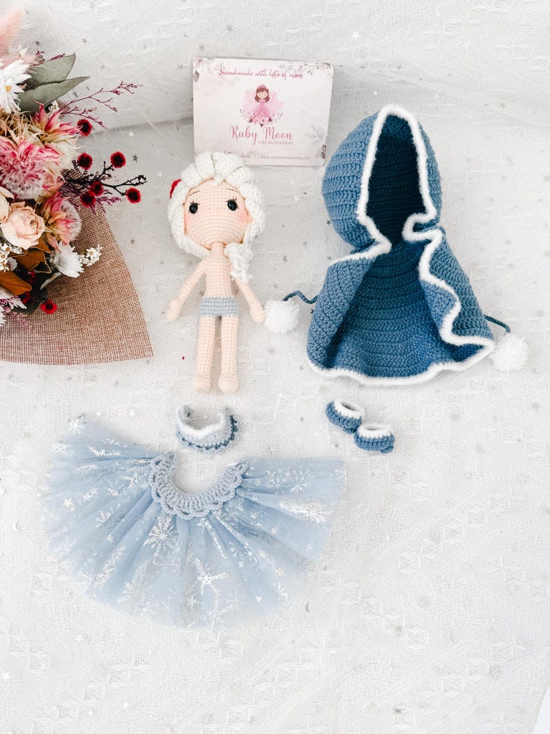 May include: A handmade crochet doll with a white hair and a blue outfit, including a hooded cape, tutu skirt, and shoes. The doll has a light skin tone and is wearing a light blue shorts. The outfit is decorated with white accents and snowflake patterns.
