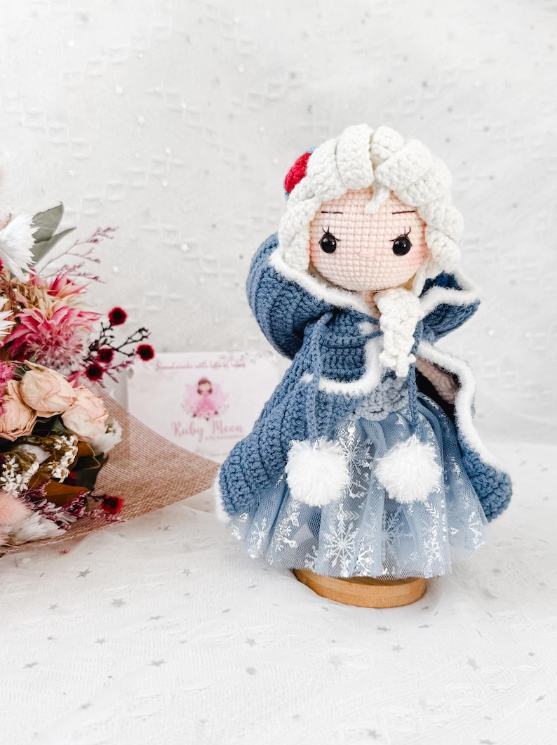 May include: A handmade crochet doll dressed in a blue coat with white trim and a light blue dress with snowflake accents. The doll has white hair and a red accent on the hat. The doll is displayed on a wooden stand.