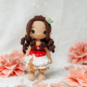May include: A handmade crochet doll with brown curly hair, a tan face, and black eyes. The doll wears a red and white top and a white skirt. A pink and green flower adorns the doll's hair. The doll is standing in front of pink flowers and petals.