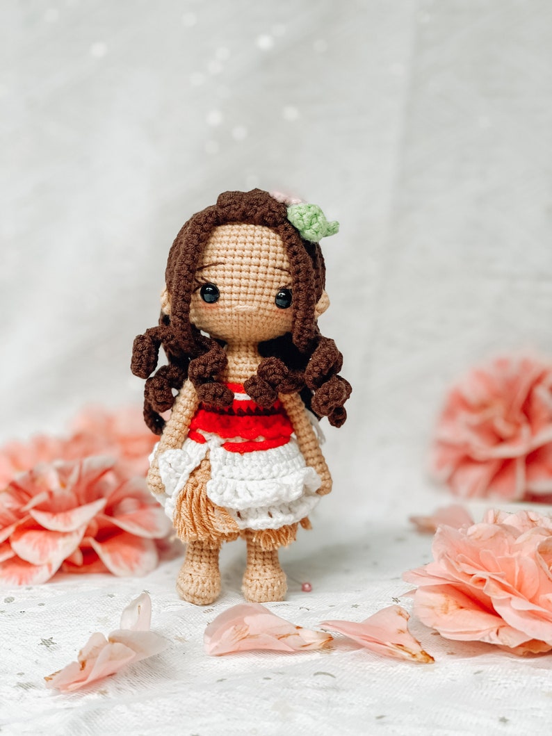 May include: A handmade crochet doll with brown curly hair, a tan face, and black eyes. The doll wears a red and white top and a white skirt. A pink and green flower adorns the doll's hair. The doll is standing in front of pink flowers and petals.