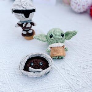 May include: Crocheted Star Wars themed toys, including a Mandalorian figure, a Grogu doll, and a creature in a pod. The figures are in shades of gray, brown, and green, with black accents. The toys are handmade and sit on a white textured surface.