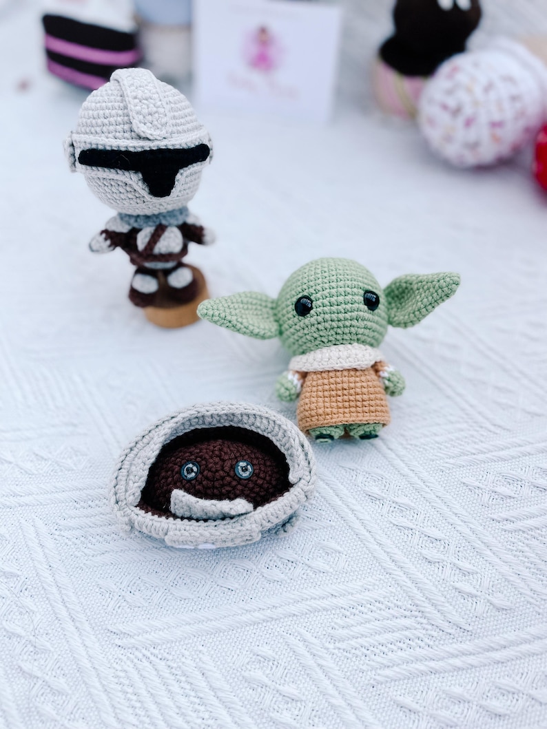 May include: Crocheted Star Wars themed toys, including a Mandalorian figure, a Grogu doll, and a creature in a pod. The figures are in shades of gray, brown, and green, with black accents. The toys are handmade and sit on a white textured surface.