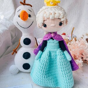 May include: Crocheted dolls of a snowman and a princess. The snowman is white with an orange nose and black eyes and buttons. The princess wears a blue dress, purple cape, and a yellow crown. The dolls are handmade.