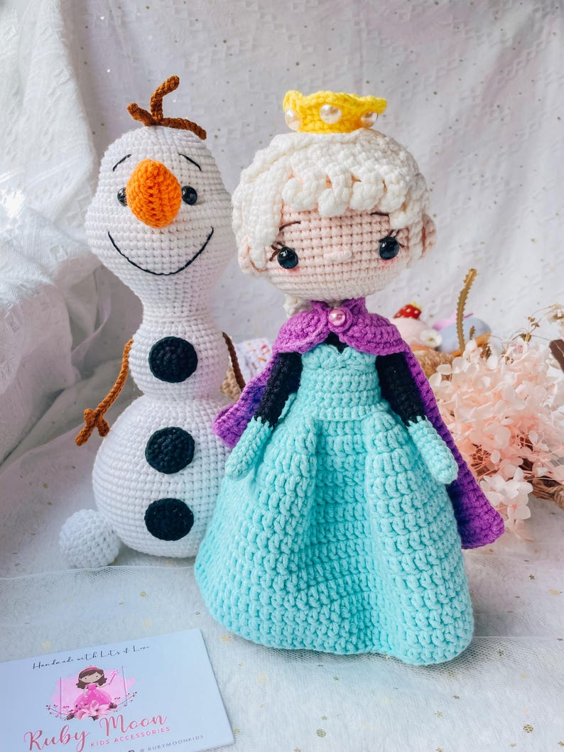 May include: Crocheted dolls of a snowman and a princess. The snowman is white with an orange nose and black eyes and buttons. The princess wears a blue dress, purple cape, and a yellow crown. The dolls are handmade.
