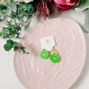 K&ouml;nnte beinhalten: Zwei gr&uuml;ne Haarklammern in Kiwi-Form auf einem rosa Teller mit Blumenmuster. Die Klammern befinden sich auf einem wei&szlig;en Etikett mit der Aufschrift "Baby Bloom".