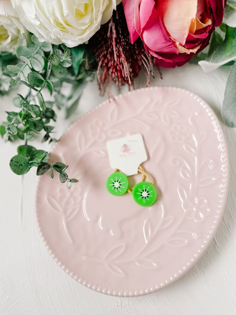 K&ouml;nnte beinhalten: Zwei gr&uuml;ne Haarklammern in Kiwi-Form auf einem rosa Teller mit Blumenmuster. Die Klammern befinden sich auf einem wei&szlig;en Etikett mit der Aufschrift "Baby Bloom".