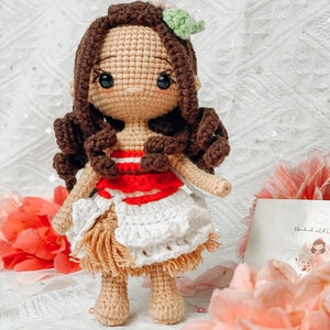 May include: A handmade crochet doll with brown hair and a floral hair accessory. The doll wears a red and white top, a white skirt, and a tan fringe. The doll is standing in front of pink flowers.