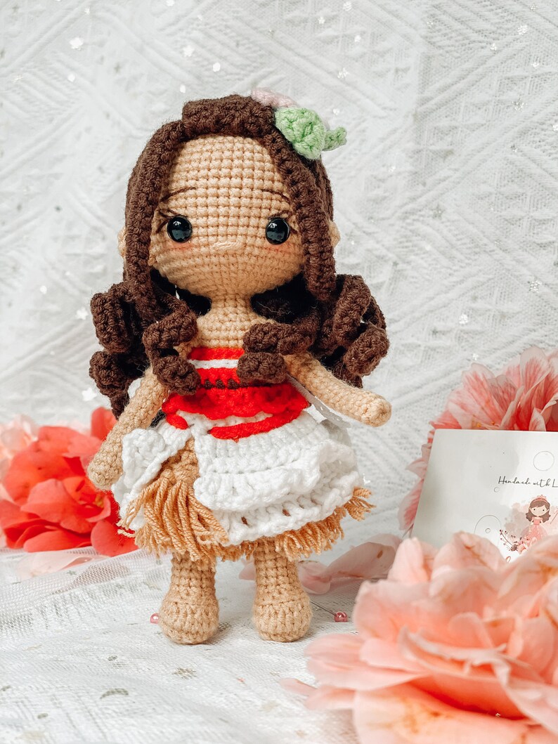 May include: A handmade crochet doll with brown hair and a floral hair accessory. The doll wears a red and white top, a white skirt, and a tan fringe. The doll is standing in front of pink flowers.