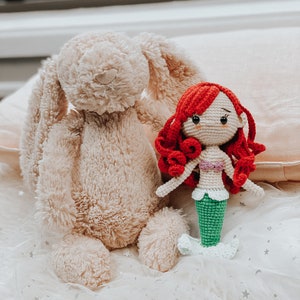 May include: A crocheted mermaid doll with red hair and a green tail sits next to a plush beige bunny on a white tulle fabric with silver stars.
