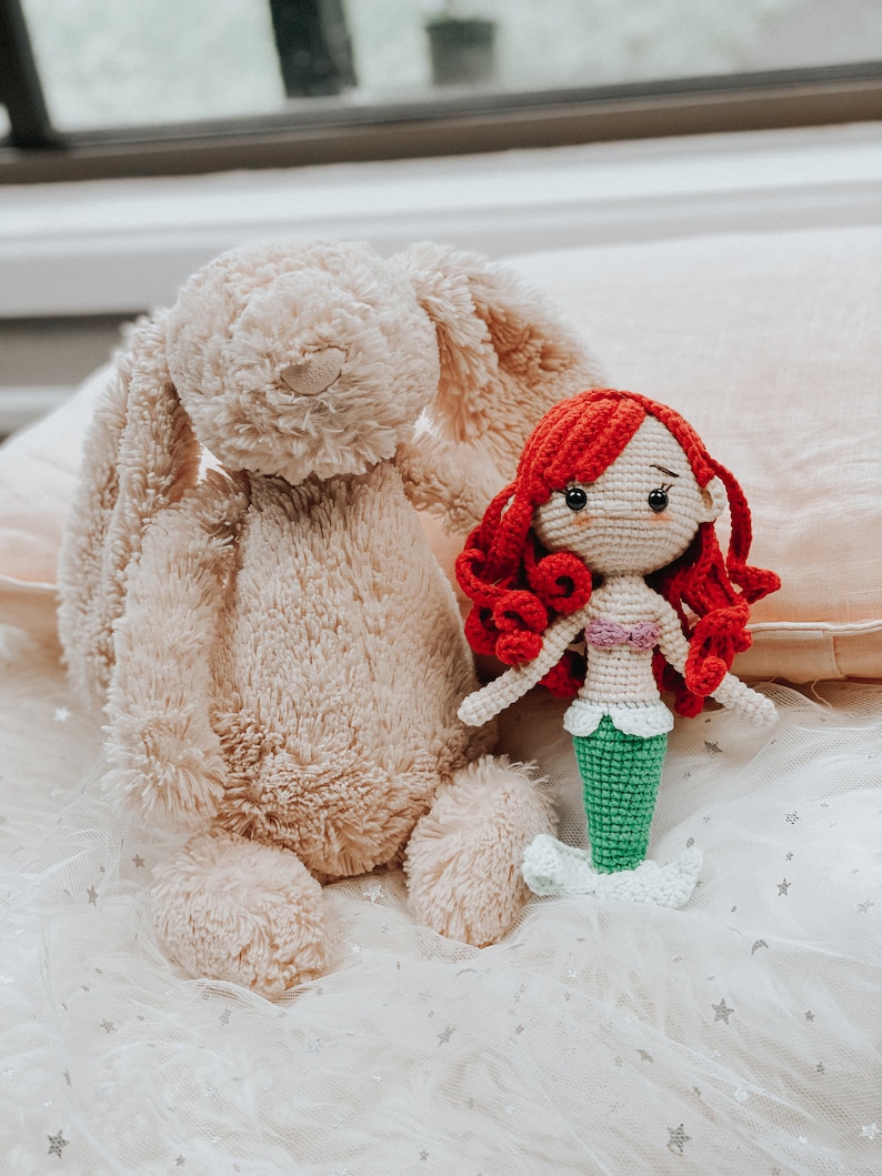 May include: A crocheted mermaid doll with red hair and a green tail sits next to a plush beige bunny on a white tulle fabric with silver stars.