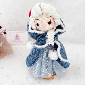 May include: A handmade crochet doll with a light skin tone, wearing a blue coat with white trim and a light blue dress with snowflake details. The doll has white hair and a red accent on the head. The doll is approximately 6 inches tall.