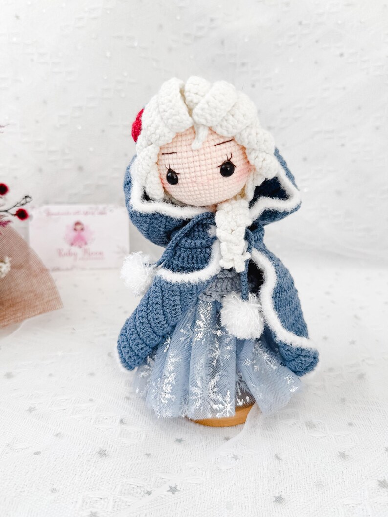 May include: A handmade crochet doll with a light skin tone, wearing a blue coat with white trim and a light blue dress with snowflake details. The doll has white hair and a red accent on the head. The doll is approximately 6 inches tall.