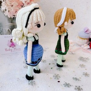 Puede incluir: Dos mu&ntilde;ecas de crochet, una con cabello blanco y un vestido azul, la otra con cabello marr&oacute;n y un vestido verde. Las mu&ntilde;ecas est&aacute;n de pie sobre una superficie blanca con copos de nieve plateados.