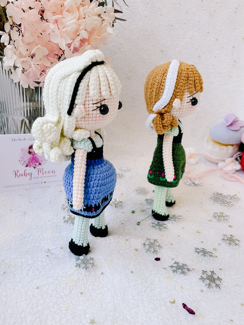 Puede incluir: Dos mu&ntilde;ecas de crochet, una con cabello blanco y un vestido azul, la otra con cabello marr&oacute;n y un vestido verde. Las mu&ntilde;ecas est&aacute;n de pie sobre una superficie blanca con copos de nieve plateados.