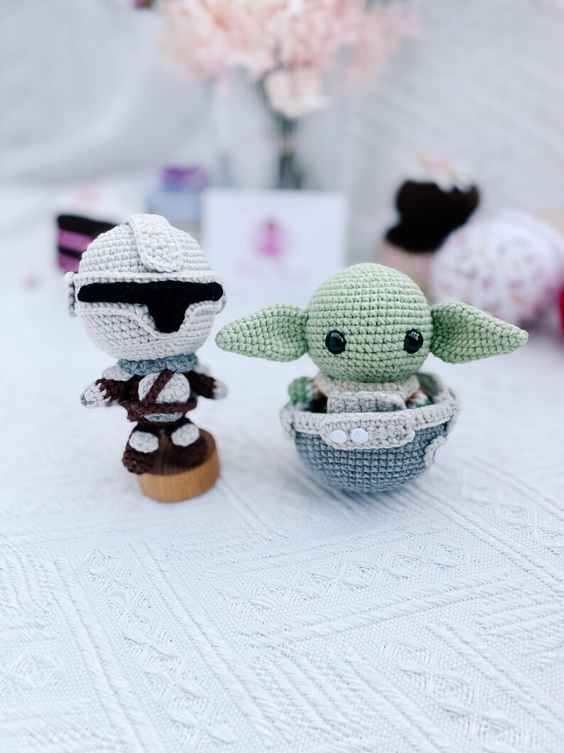 May include: Two crocheted figures: a Stormtrooper and a Grogu (Baby Yoda). The Stormtrooper is in a white helmet and armor, with a black visor. Grogu is green, with large ears, sitting in a gray and blue crocheted pod. Both figures are on a white surface.