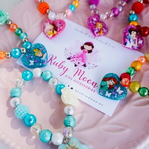 May include: A set of colorful beaded bracelets with princess-themed charms. Each bracelet features a different princess character, including Ariel, Cinderella, and Elsa. The bracelets are made with a variety of beads, including clear, iridescent, and glitter beads. The bracelets are packaged on a pink plate with a business card that reads "Ruby Moon Kids Accessories" and "@rubymoonkids".