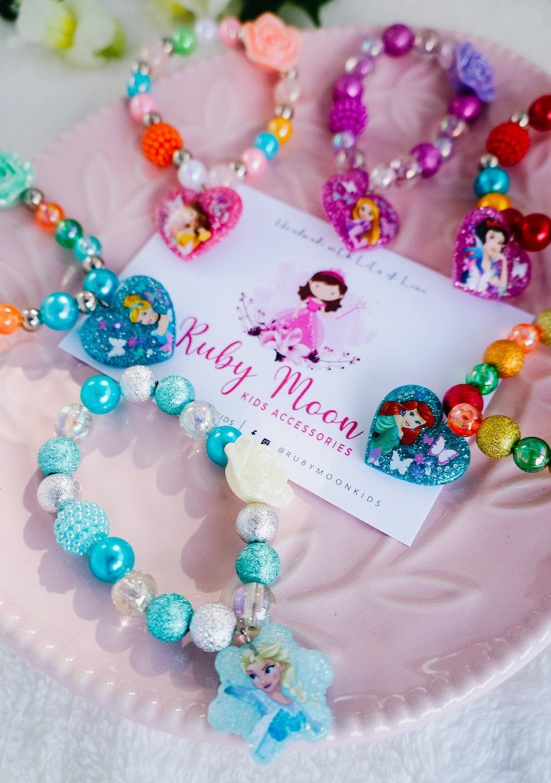 May include: A set of colorful beaded bracelets with princess-themed charms. Each bracelet features a different princess character, including Ariel, Cinderella, and Elsa. The bracelets are made with a variety of beads, including clear, iridescent, and glitter beads. The bracelets are packaged on a pink plate with a business card that reads "Ruby Moon Kids Accessories" and "@rubymoonkids".