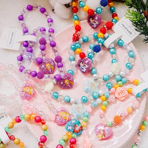 Pode incluir: Uma cole&ccedil;&atilde;o de colares e pulseiras de contas coloridas com personagens de princesas Disney. Cada pe&ccedil;a tem um pingente em forma de cora&ccedil;&atilde;o com uma princesa diferente, incluindo Rapunzel, Cinderela, Ariel e Bela. Os colares e as pulseiras s&atilde;o feitos com contas de v&aacute;rias cores, incluindo roxo, azul, rosa, amarelo e verde. O texto "Hand made" &eacute; vis&iacute;vel em duas das etiquetas.