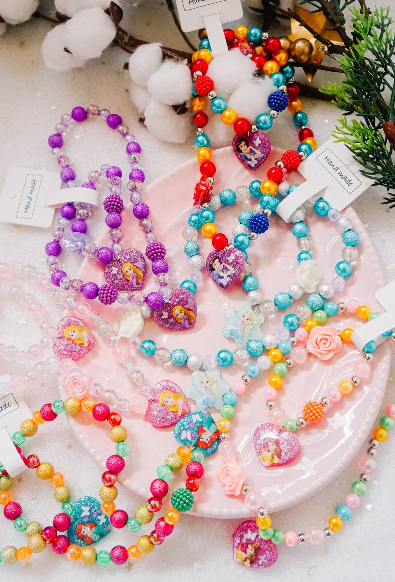 Pode incluir: Uma cole&ccedil;&atilde;o de colares e pulseiras de contas coloridas com personagens de princesas Disney. Cada pe&ccedil;a tem um pingente em forma de cora&ccedil;&atilde;o com uma princesa diferente, incluindo Rapunzel, Cinderela, Ariel e Bela. Os colares e as pulseiras s&atilde;o feitos com contas de v&aacute;rias cores, incluindo roxo, azul, rosa, amarelo e verde. O texto "Hand made" &eacute; vis&iacute;vel em duas das etiquetas.